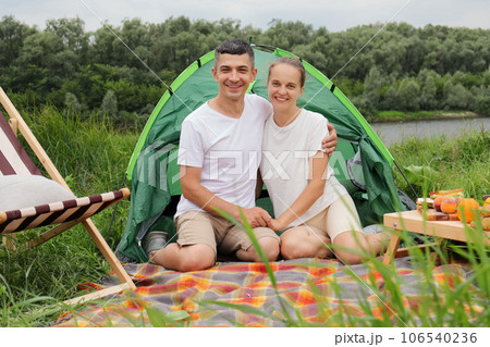 Smiling happy optimistic woman and man wearing casual clothing sitting at tent near the river looking at camera having picnic enjoying spring days. 106540236