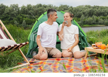 Tasty coffee on nature. Joyful smiling woman and man wearing casual clothing sitting at tent near the river, drinking coffee, enjoying aromat, resting, relaxing. 106540238