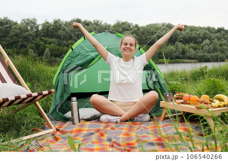 Cheerful woman wearing white t shirt sitting near the tent with crossed legs, raised arms, rejoicing active weekend on nature, looking at camera. Cheerful woman wearing white t shirt sitting near the tent with crossed legs, raised arms, rejoicing active weekend on nature, looking at camera. 106540266
