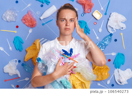 Sad unhappy woman wearing white shirt isolated on blue wall with litter around cleaning territory from rubbish looking at camera with displeased expression. Sad unhappy woman wearing white shirt isolated on blue wall with litter around cleaning territory from rubbish looking at camera with displeased expression. 106540273