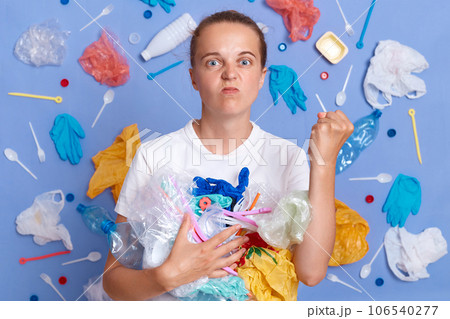 Volunteers clean up garbage. Reuse materials to reduce waste. Angry woman wearing white shirt isolated on blue wall with litter around with rubbish showing raised fist. 106540277