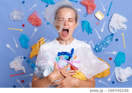 Despair sad unhappy woman wearing white shirt isolated on blue wall with rubbish around holding trash cleaning parks screaming from shock and sorrow environmental pollution. Despair sad unhappy woman wearing white shirt isolated on blue wall with rubbish around holding trash cleaning parks screaming from shock and sorrow environmental pollution. 106540288