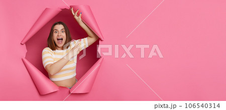 Positive optimistic brunette woman wearing striped top looking through ripped pink paper hole pointing away at copy space for promotion advertisement area. 106540314