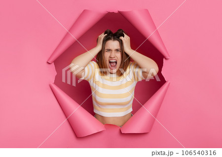 Irritated frustrated brunette woman wearing striped top looking through ripped pink paper hole being in panic pulling her hair screaming with anger. Irritated frustrated brunette woman wearing striped top looking through ripped pink paper hole being in panic pulling her hair screaming with anger. 106540316