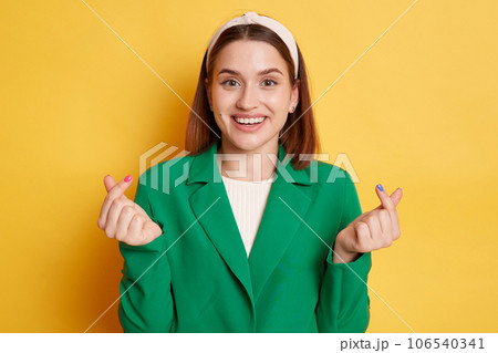 Excited amazed woman wearing green jacket posing isolated over yellow background standing showing korean love gesture expressing love. Excited amazed woman wearing green jacket posing isolated over yellow background standing showing korean love gesture expressing love. 106540341