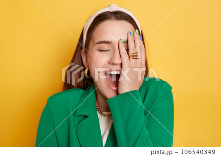 Positive laughing woman wearing green jacket standing isolated over yellow background showing facepalm covering half of face with hand closed eyes. 106540349