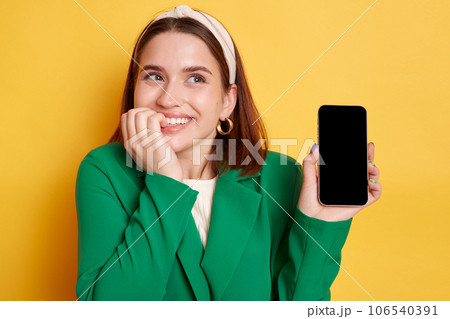Dreaming smiling satisfied woman wearing green jacket posing isolated over yellow background showing cell phone advertisement empty space for promotion. Dreaming smiling satisfied woman wearing green jacket posing isolated over yellow background showing cell phone advertisement empty space for promotion. 106540391