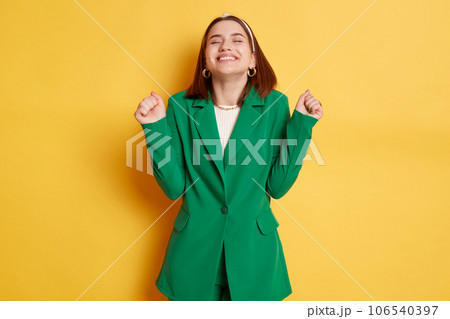 Extremely happy woman wearing green jacket standing isolated over yellow background closed her eyes clenched fists celebrating her success. Extremely happy woman wearing green jacket standing isolated over yellow background closed her eyes clenched fists celebrating her success. 106540397