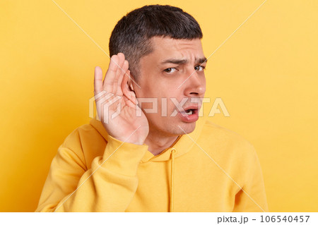 Confused man wearing casual hoodie standing with hands near ear trying to listen silent talking posing isolated over yellow background. Confused man wearing casual hoodie standing with hands near ear trying to listen silent talking posing isolated over yellow background. 106540457
