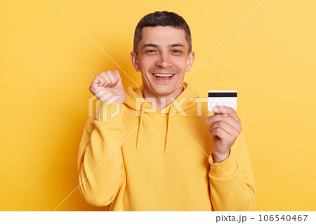 Online shopping. Banking paying. Delighted cheerful man wearing casual hoodie earning big sum of money clenched fist celebrating succes posing isolated over yellow background. 106540467