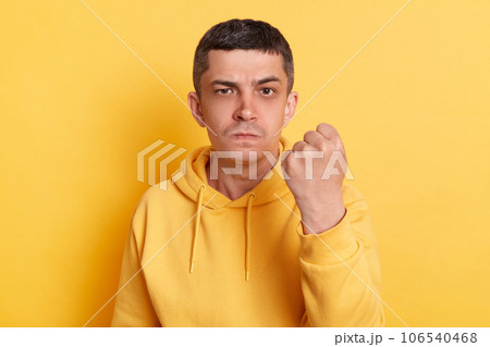 Aggressive guy. Furious quarrel. Serious strict man wearing casual hoodie showing clenched fist arguing with wife looking at camera posing isolated over yellow background. 106540468
