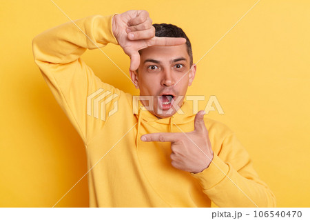 Shocked photographer. Surprised astonished man wearing casual hoodie making frame of fingers looking at camera with open mouth posing isolated over yellow background. Shocked photographer. Surprised astonished man wearing casual hoodie making frame of fingers looking at camera with open mouth posing isolated over yellow background. 106540470