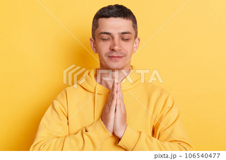 Meditation. Practicing yoga. Calm relaxed man wearing casual hoodie keeps palms together closed eyes doing yoga for relaxing posing isolated over yellow background. Meditation. Practicing yoga. Calm relaxed man wearing casual hoodie keeps palms together closed eyes doing yoga for relaxing posing isolated over yellow background. 106540477