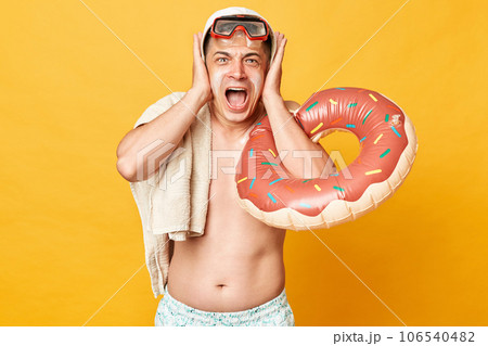 Irritated adult man wearing shorts swimsuit snorkeling goggles holding donut rubber ring and bag isolated on yellow background screaming being depressed covering ears with mouth. Irritated adult man wearing shorts swimsuit snorkeling goggles holding donut rubber ring and bag isolated on yellow background screaming being depressed covering ears with mouth. 106540482