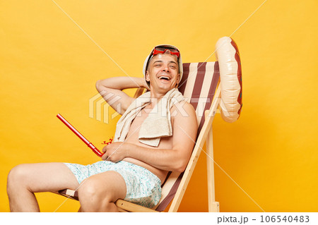 Relaxed satisfied smiling shirtless man sitting on deck chair isolated over yellow background enjoying his summer vacation laughing happily. 106540483