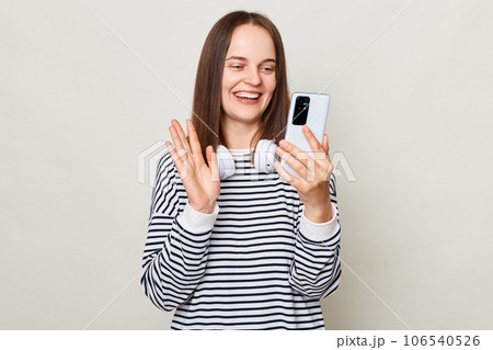 Cheerful joyful brown haired adult woman wearing striped shirt standing isolated over gray background holding smart phone having video call waving hand. Cheerful joyful brown haired adult woman wearing striped shirt standing isolated over gray background holding smart phone having video call waving hand. 106540526