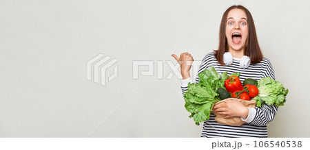 Amazed excited brown haired young woman embraces bouquet of fresh vegetables wearing striped casual shirt isolated over gray background pointing at copy space for promotion. 106540538