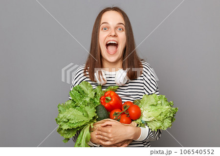 Grocery shopping for healthy eating. Shopping at the vegetarian market. Amazed excited woman embracing fresh vegetables returns from supermarket wearing striped  shirt isolated over gray background 106540552
