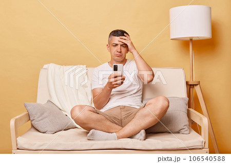 Sad unhappy brunette adult man wearing casual clothing sitting on sofa against beige wall using smartphone reading bad news holding hand on his forehead. Sad unhappy brunette adult man wearing casual clothing sitting on sofa against beige wall using smartphone reading bad news holding hand on his forehead. 106540588
