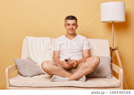 Handsome smiling charming brunette adult man wearing casual clothing sitting on sofa against beige wall looking at camera with relaxed satisfied look. 106540589
