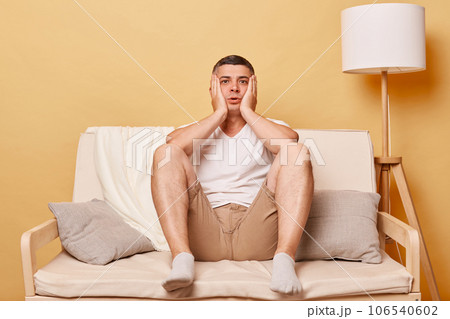 Shocked scared brunette young man wearing casual white t shirt sitting on sofa against beige wall keeps hands on cheeks looking at camera with big eyes full of fear. Shocked scared brunette young man wearing casual white t shirt sitting on sofa against beige wall keeps hands on cheeks looking at camera with big eyes full of fear. 106540602