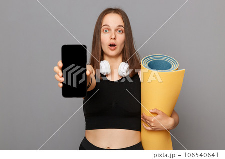 Astonished woman wearing black sportswear holding yoga mat standing isolated over gray background looking at camera with big eyes showing phone with empty screen. 106540641