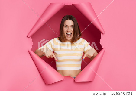 Excited positive attractive woman wearing striped shirt breaking through pink paper hole pointing down at copy space for promotion, advertisement area. 106540701