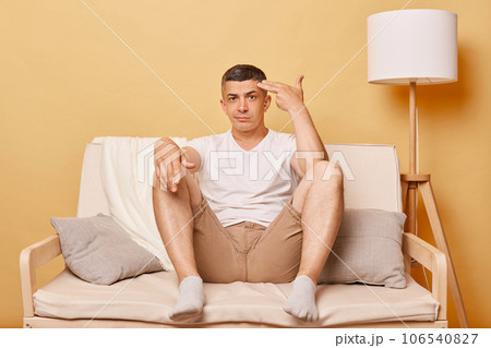 Stressed depressed brunette adult man wearing casual clothing sitting on sofa against beige wall making suicide gesture with fingers gun, looking at camera with displeased expression. Stressed depressed brunette adult man wearing casual clothing sitting on sofa against beige wall making suicide gesture with fingers gun, looking at camera with displeased expression. 106540827