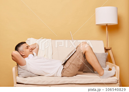 Smiling handsome man lying on sofa at home watching movie or film on laptop in the living room having satisfied expression being calm and relaxed keeps hands behind head. 106540829