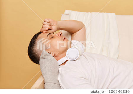 Adult man lies on the couch at home against beige background suffering headache or symptoms of the disease, being tired exhausted fellign stress and fatigue. Adult man lies on the couch at home against beige background suffering headache or symptoms of the disease, being tired exhausted fellign stress and fatigue. 106540830