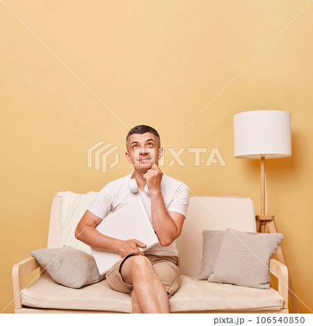 Pensive thoughtful adult man wearing white t-shirt and shorts sitting on sofa with closed laptop against beige wall looking upwards at advertisement area copy space for promotion. Pensive thoughtful adult man wearing white t-shirt and shorts sitting on sofa with closed laptop against beige wall looking upwards at advertisement area copy space for promotion. 106540850