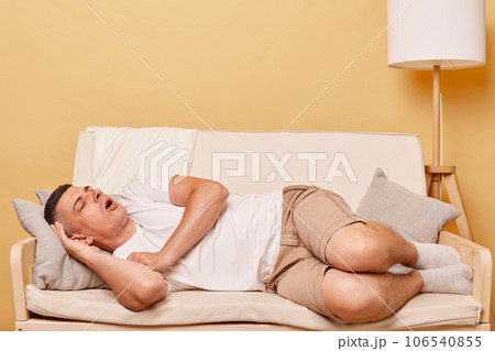 Brunette guy sleeping on the couch having funny expression lying with open mouth resting while having free time home napping at weekend. Brunette guy sleeping on the couch having funny expression lying with open mouth resting while having free time home napping at weekend. 106540855