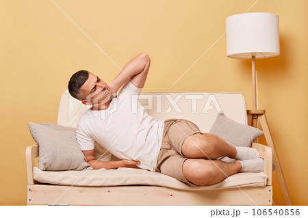 Handsome unhealthy man wearing white t-shirt suffering backache feeling strong pain in neck massaging grimacing frowning face lying on sofa against beige wall. 106540856