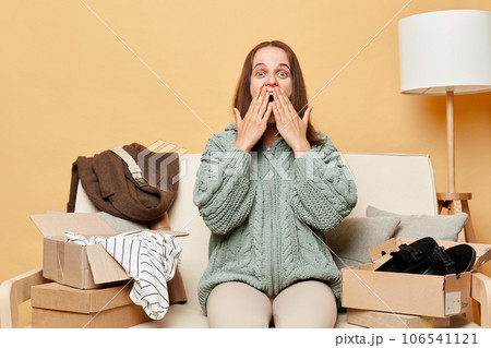 Shocked surprised woman sitting on sofa among boxes with clothing against beige wall relocating to new apartment unpacking her belongings looking at camera saying wow Shocked surprised woman sitting on sofa among boxes with clothing against beige wall relocating to new apartment unpacking her belongings looking at camera saying wow 106541121