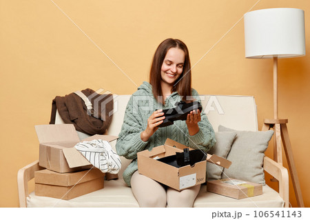 Attractive happy positive woman sitting on sofa among boxes with clothing against beige wall buying new shoes admiring unpacking package from shop delivery. Attractive happy positive woman sitting on sofa among boxes with clothing against beige wall buying new shoes admiring unpacking package from shop delivery. 106541143