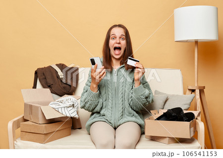 Excited amazed happy woman sitting on sofa among boxes with clothing against beige wall posing with mobile phone and credit card getting cashback, rejoicing. 106541153
