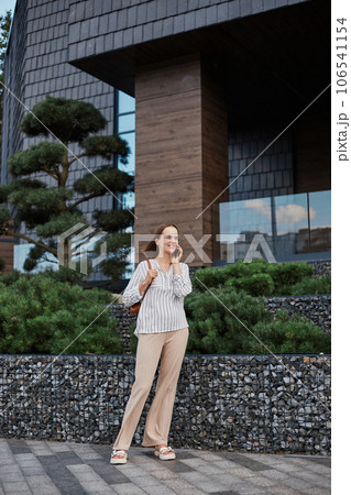 Full length portrait of attractive woman in elegant blouse and trousers standing in street and talking on mobile phone looking smiling away having business conversation. Full length portrait of attractive woman in elegant blouse and trousers standing in street and talking on mobile phone looking smiling away having business conversation. 106541154
