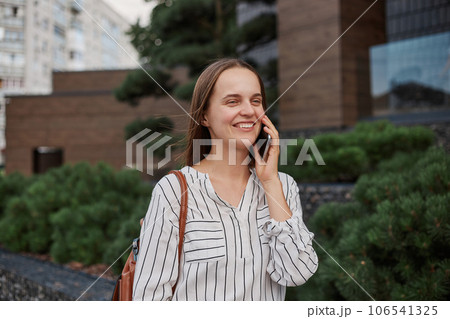 Charming smiling positive woman with pleasant appearance looking away with happy face talking on mobile phone enjoying conversation walking outside. Charming smiling positive woman with pleasant appearance looking away with happy face talking on mobile phone enjoying conversation walking outside. 106541325