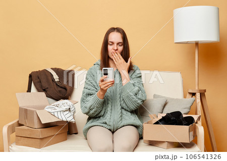 Surprised woman wearing knitted sweater with ordered clothes in boxes sitting at home on couch against beige wall using mobile phone get mistake parcel checking app. 106541326