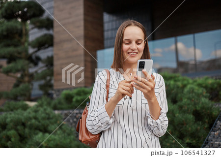 Good looking positive woman wearing elegant clothes standing with smartphone in hands expressing positive emotions reading news browsing internet. Good looking positive woman wearing elegant clothes standing with smartphone in hands expressing positive emotions reading news browsing internet. 106541327