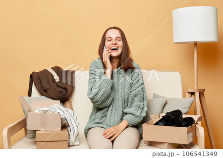 Attractive laughing woman sitting on sofa among boxes with clothing against beige wall talking on smartphone with friend saying about internet shopping. 106541349