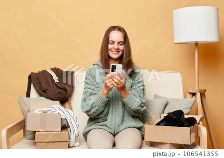 Charming cheerful woman wearing knitted sweater with ordered clothes in boxes sitting at home on couch against beige wall chatting with friends on smartphone boasting her online shopping. Charming cheerful woman wearing knitted sweater with ordered clothes in boxes sitting at home on couch against beige wall chatting with friends on smartphone boasting her online shopping. 106541355