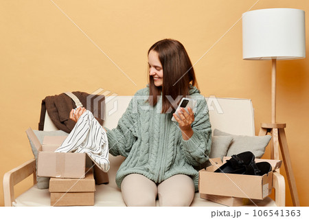 Satisfied smiling beautiful woman sitting on sofa among boxes with clothing against beige wall checking her order with mobile phone being delighted expressing positive emotions. 106541363