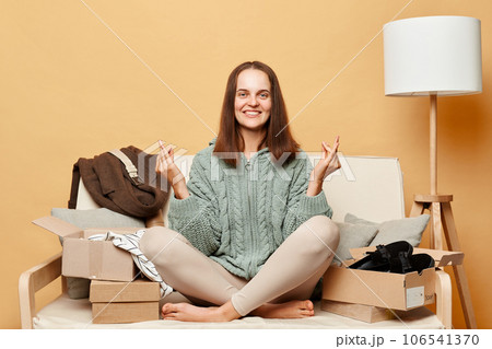 Smiling attractive woman shopaholic customer sitting on sofa unpack parcel delivery box against beige wall posing with mudra gesture practicing yoga. Smiling attractive woman shopaholic customer sitting on sofa unpack parcel delivery box against beige wall posing with mudra gesture practicing yoga. 106541370