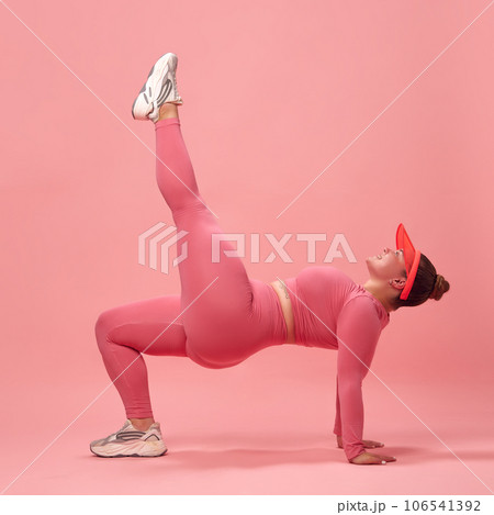 Full body side view of young overweight sporty woman in pink top and legging, training legs and back isolated studio background. Single leg bridge. 106541392