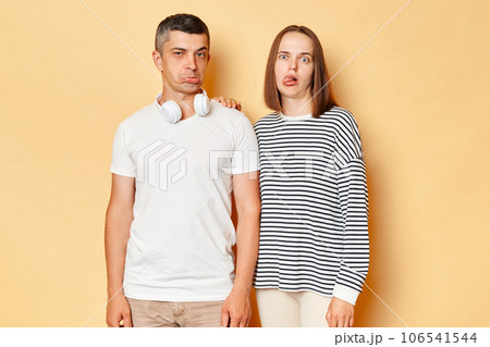 Funny childish young couple wife and husband wearing casual attires standing isolated over beige background frimacing showing tongue out and pout lips playful behavior. 106541544