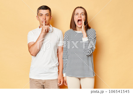 Caucasian woman and man wearing casual outfits standing isolated over beige background, guy showing shh gesture, female screaming loud with hand near mouth. 106541545