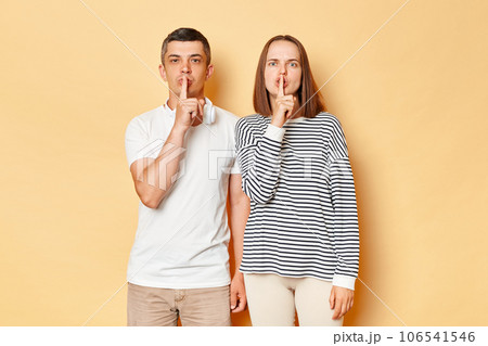 Serious strict young couple friends man woman in casual clothing isolated over beige background keeping fingers near mouths keep secret looking at cmaera with bossy expressions. 106541546