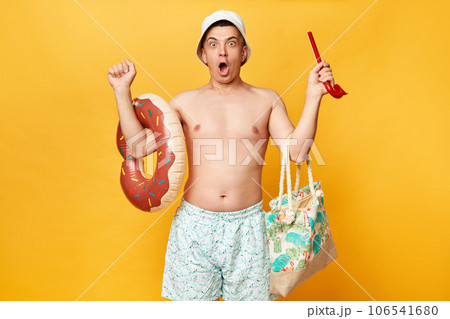 Amazed man wearing shorts swimsuit and panama holding donut rubber ring isolated on yellow background looking at camera with big eyes keeps mouth open raised arms. 106541680