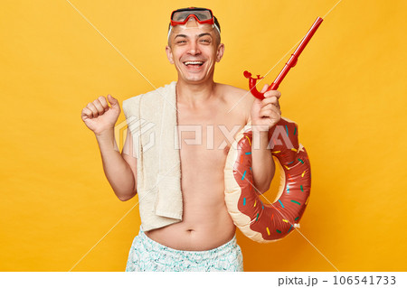 Happy cheerful man in diving goggles and snorkel holding donut rubber ring isolated on yellow background rejoicing his simmer activity clenched fists smiling. 106541733